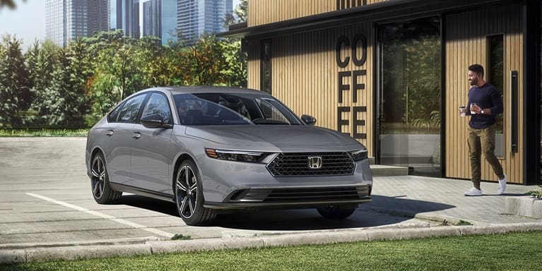 2026 Honda Accord 3/4 front view parked out in front of a cafe in the early morning with a person walking back to the car with two coffees in his hands.